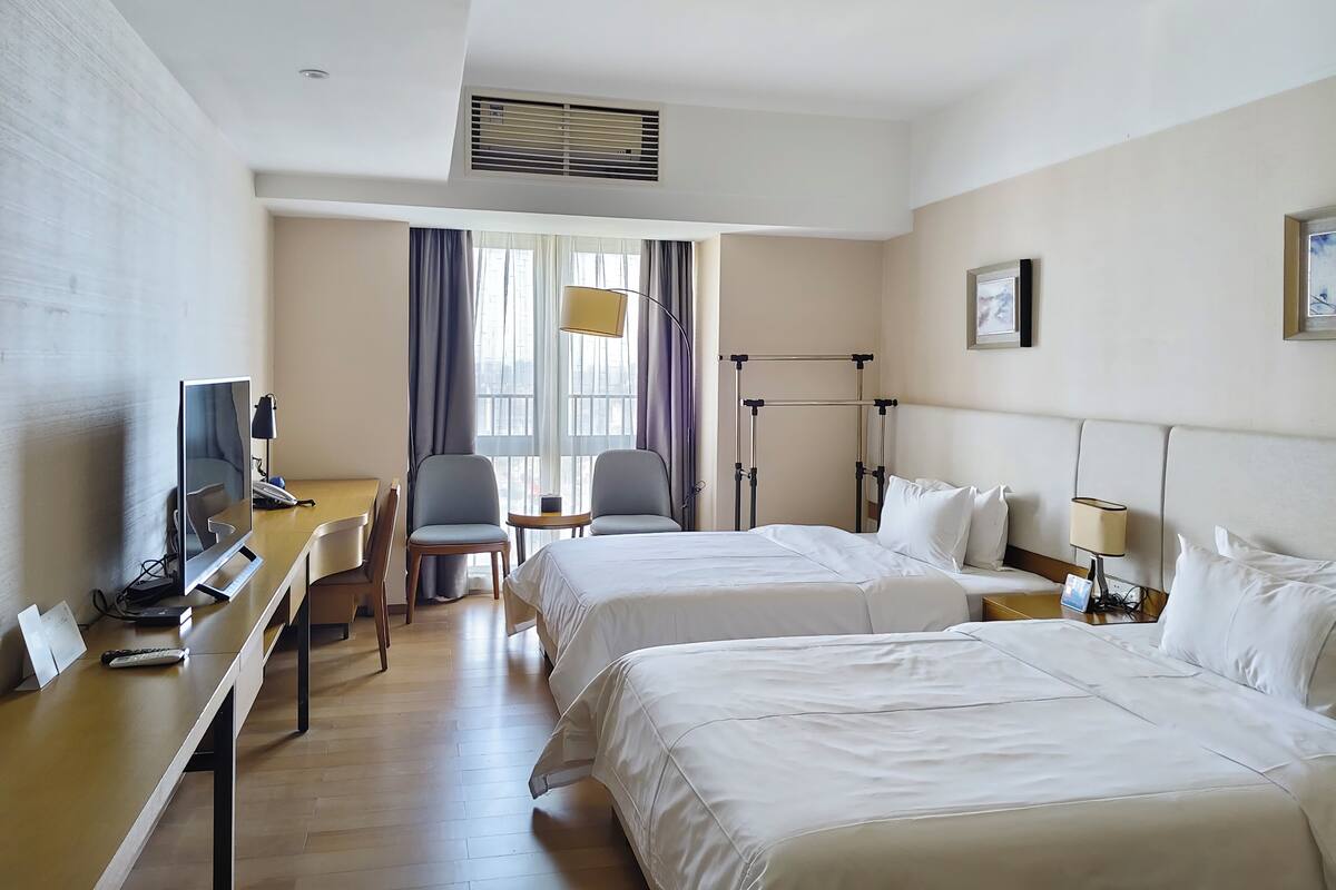 A hotel room with two beds, a desk with a computer, a chair, a lamp, and a window with curtains.