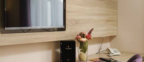 A hotel room with a mounted TV, a desk with a vase of flowers, a phone, and a book.