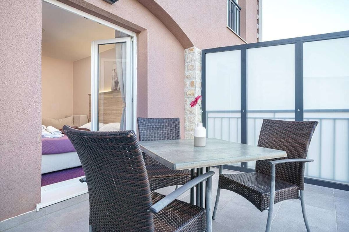 An outdoor balcony with a table and chairs, a sliding glass door leading to a room, and a flower vase on the table.