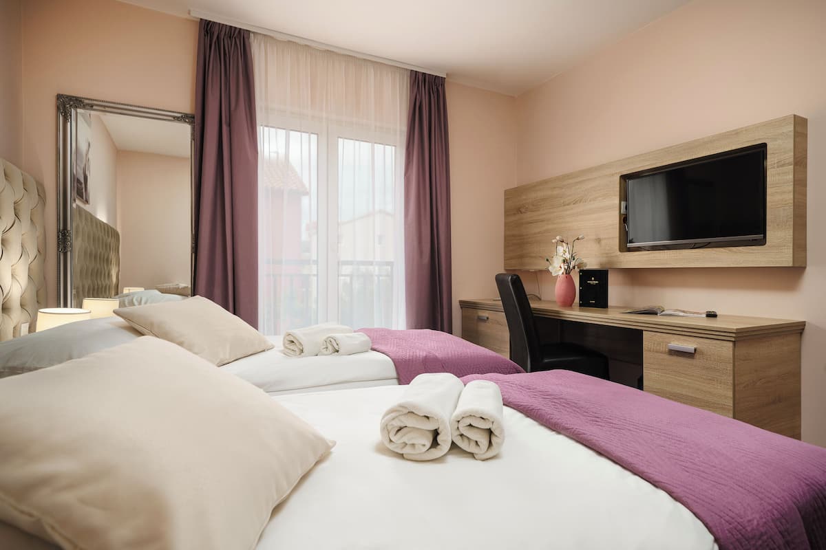 A hotel room with two beds, a desk, a chair, a TV, and a large window with curtains.