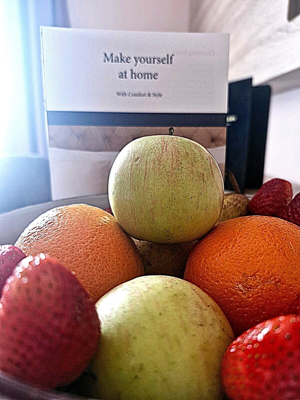 Un tazón de fruta con manzanas y naranjas, y un letrero al fondo que dice 'Hazte sentir como en casa'.