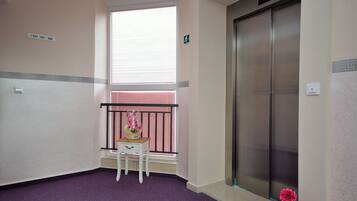 Een hoekappartement met een paars tapijt, een witte stoel, een klein tafeltje met een bloevas, een raam met jaloezieën en een lift.