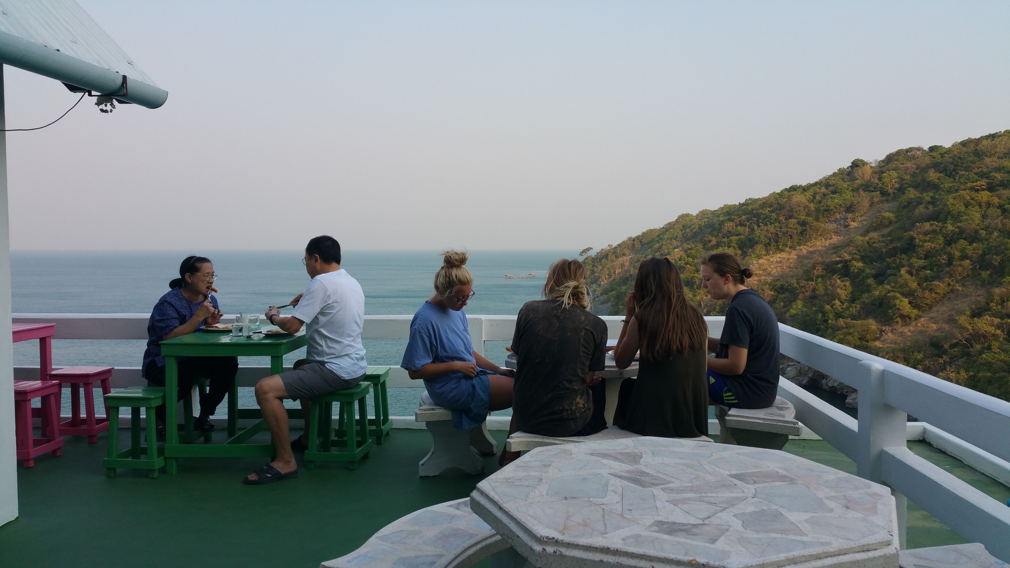 Um grupo de pessoas jantando em uma mesa ao ar livre, com vista para o mar e uma paisagem montanhosa.