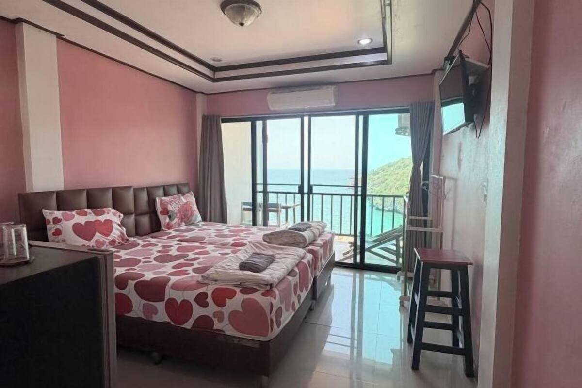 A bedroom with a large bed, a desk, a chair, and a balcony with a view.