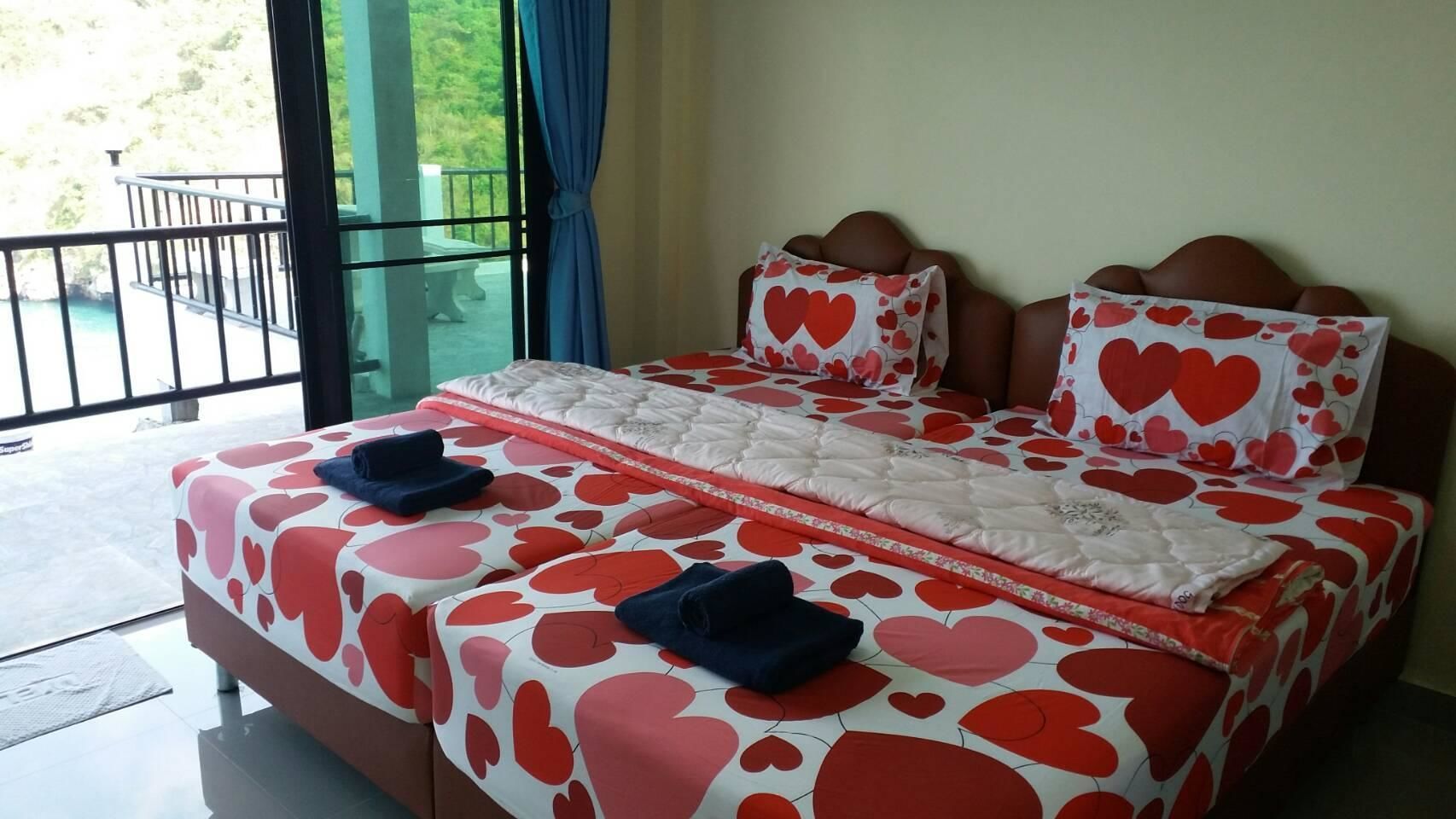 Cama doble con ropa de cama estampada con motivos de corazón, vista a zonas verdes a través de un balcón y una puerta de cristal.
