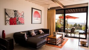 A modern living room with a black leather sofa, a coffee table, and a large window overlooking a garden. - Vista Real Villas (Corregidora)
