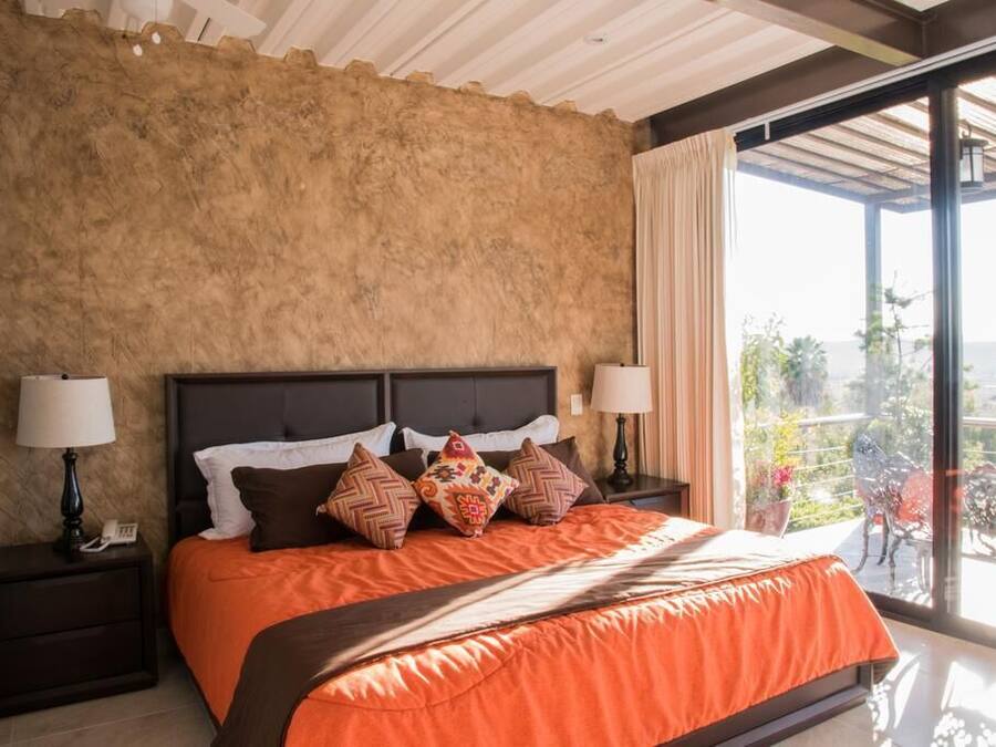 A bedroom with a large bed, two bedside tables with lamps, a view of the outdoors through a large window, and a balcony with chairs.