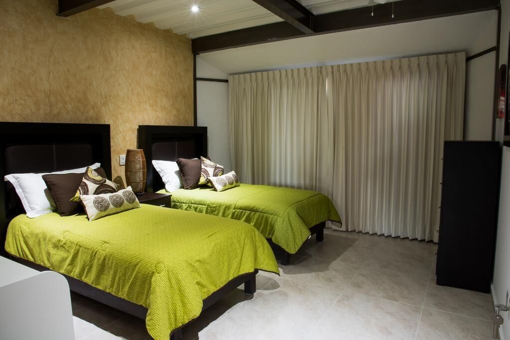 A hotel room with two beds, each with a green bedspread and multiple pillows. The room has a neutral color scheme with beige walls and a large window with curtains.