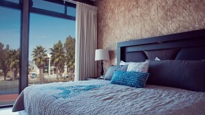 A large bed with a quilt, a nightstand with a lamp, and a view of palm trees through a large window. - Vista Real Villas (Corregidora)