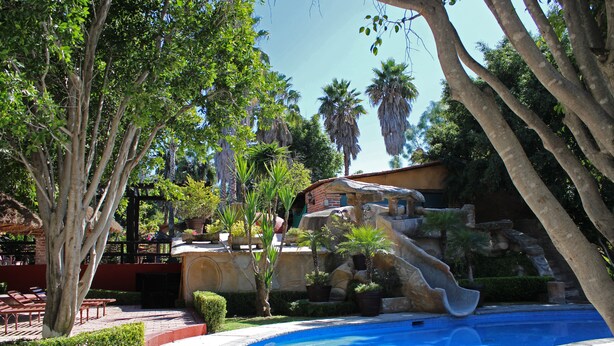 A pool area with a waterfall feature, surrounded by lush greenery and palm trees. - Vista Real Villas (Corregidora)