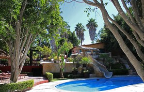 A pool area with a waterfall feature, surrounded by lush greenery and palm trees. - Vista Real Villas (Corregidora)