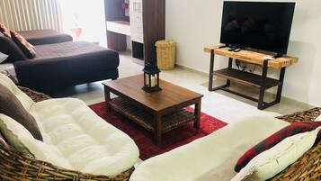 A living room with a sofa, a coffee table, a TV on a wooden stand, and wicker furniture.