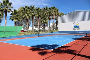 A tennis court with a blue and red surface, surrounded by palm trees and a building with a logo. - Vista Real Villas (Corregidora)