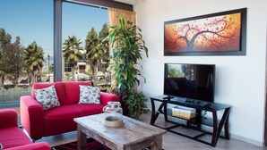 A living room with a red sofa, a wooden coffee table, a TV on a stand, and a large window with a view of palm trees and buildings. - Vista Real Villas (Corregidora)