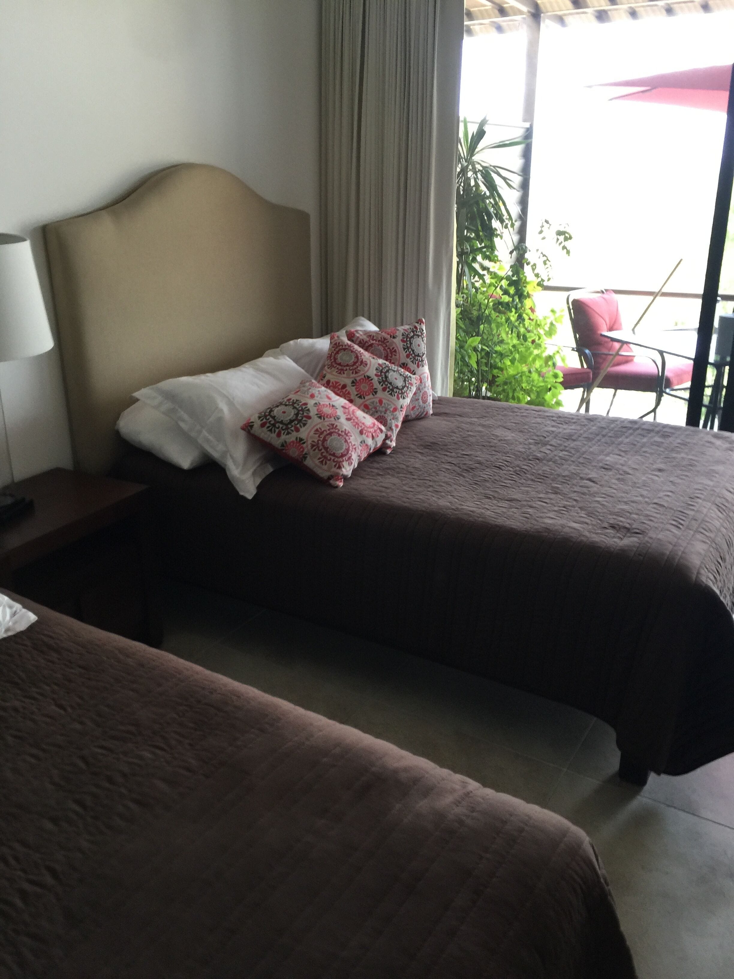 Camera d'albergo con due letti, ciascuno con copripiumino marrone e cuscini decorativi. Si nota un comodino con una lampada, una finestra con tende e vista sull'esterno.