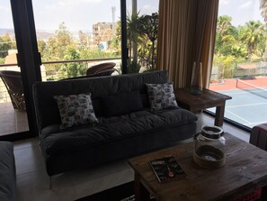 A modern living room with a dark grey sofa, two patterned cushions, a wooden coffee table with a magazine, and a view of a tennis court and greenery outside. - Vista Real Villas (Corregidora)