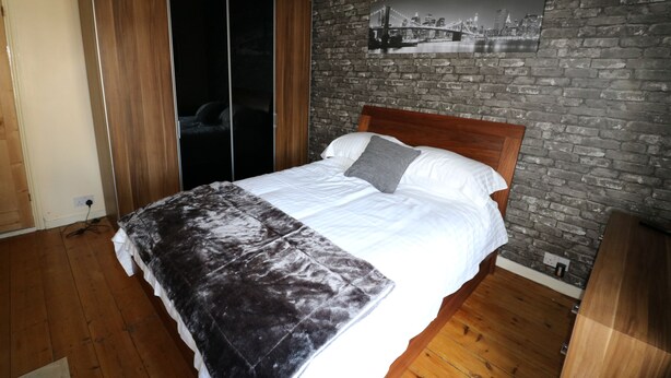 A bedroom with a wooden bed, a grey blanket, a cityscape picture on the brick wall, and a sliding wardrobe. - Victoria House (Colchester)
