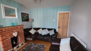 A living room with a brick fireplace, a dark brown leather sofa, and a grey rug. - Victoria House (Colchester)