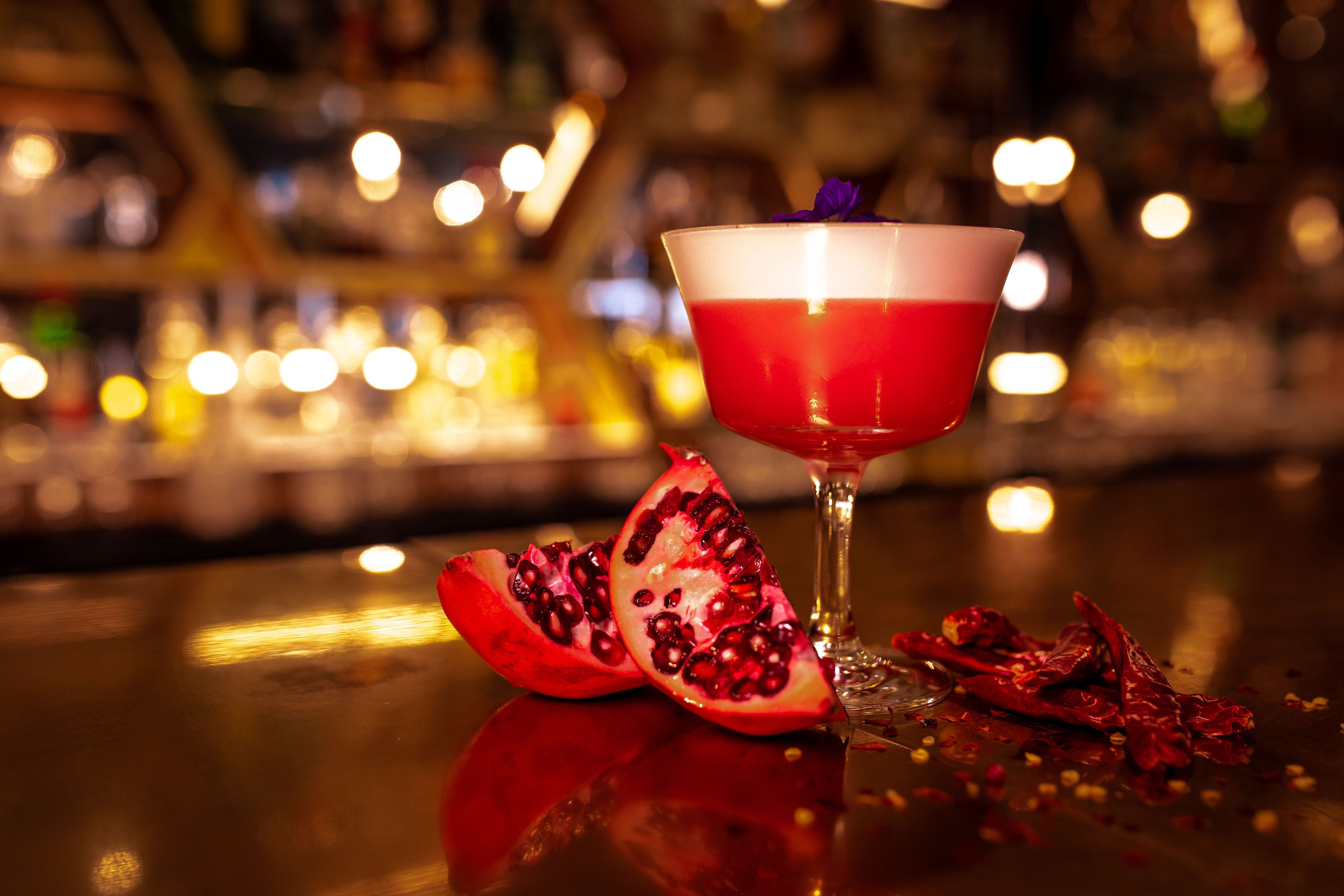 A cocktail glass with a red and white rim, a pomegranate half, and dried red chili peppers on a reflective surface.
