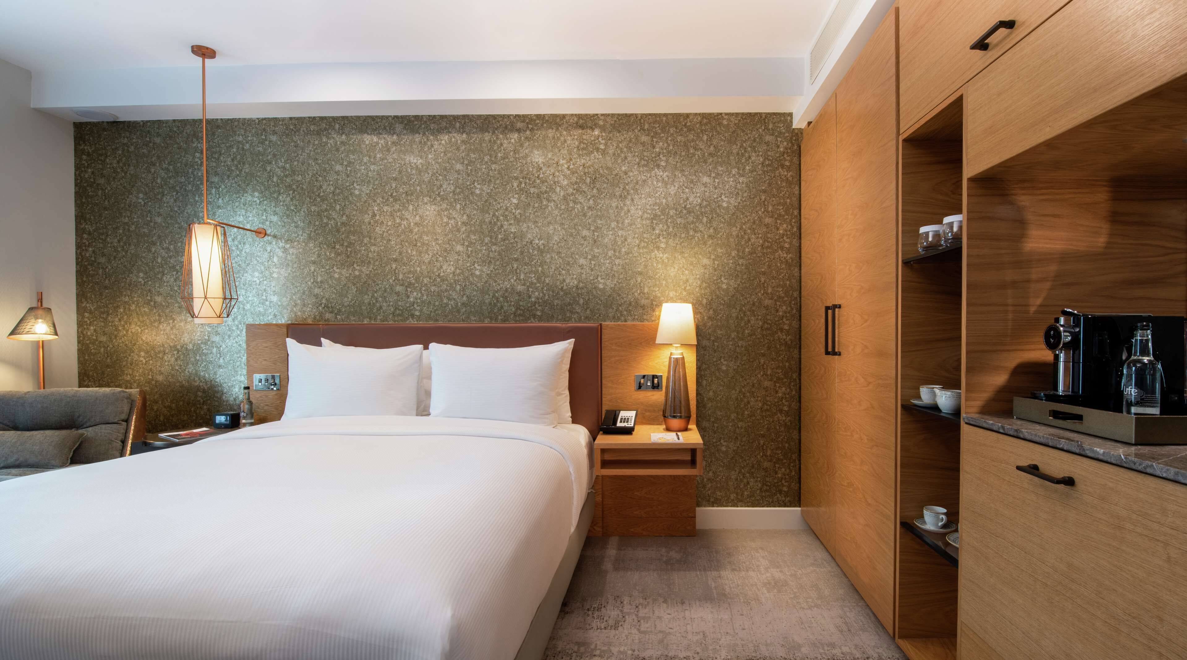 A hotel room with a large bed, a nightstand with a lamp, a wooden wardrobe, and a kitchenette area.