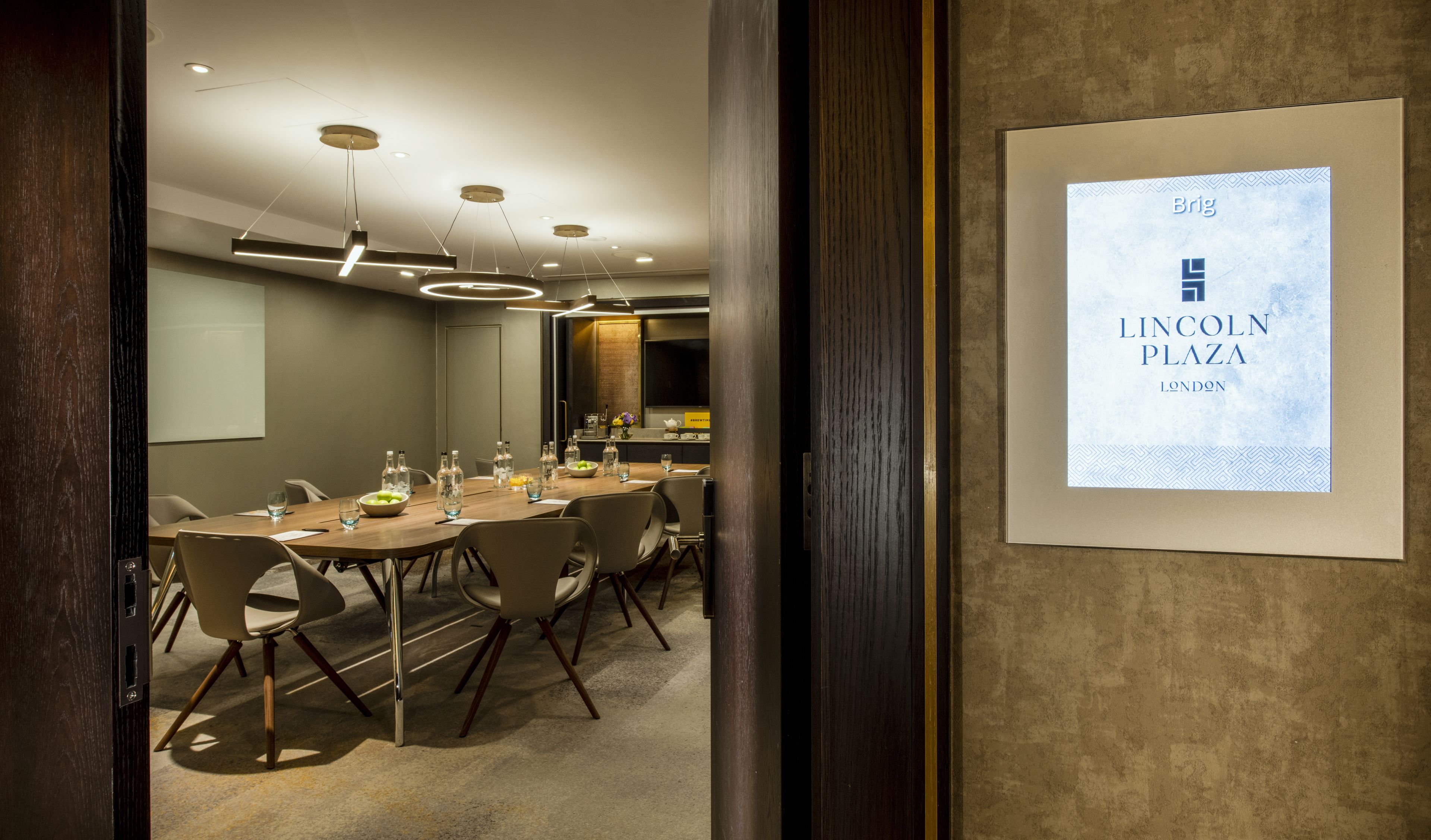 A conference room with a long table, chairs, and a projector screen displaying "Brig Lincoln Plaza London".