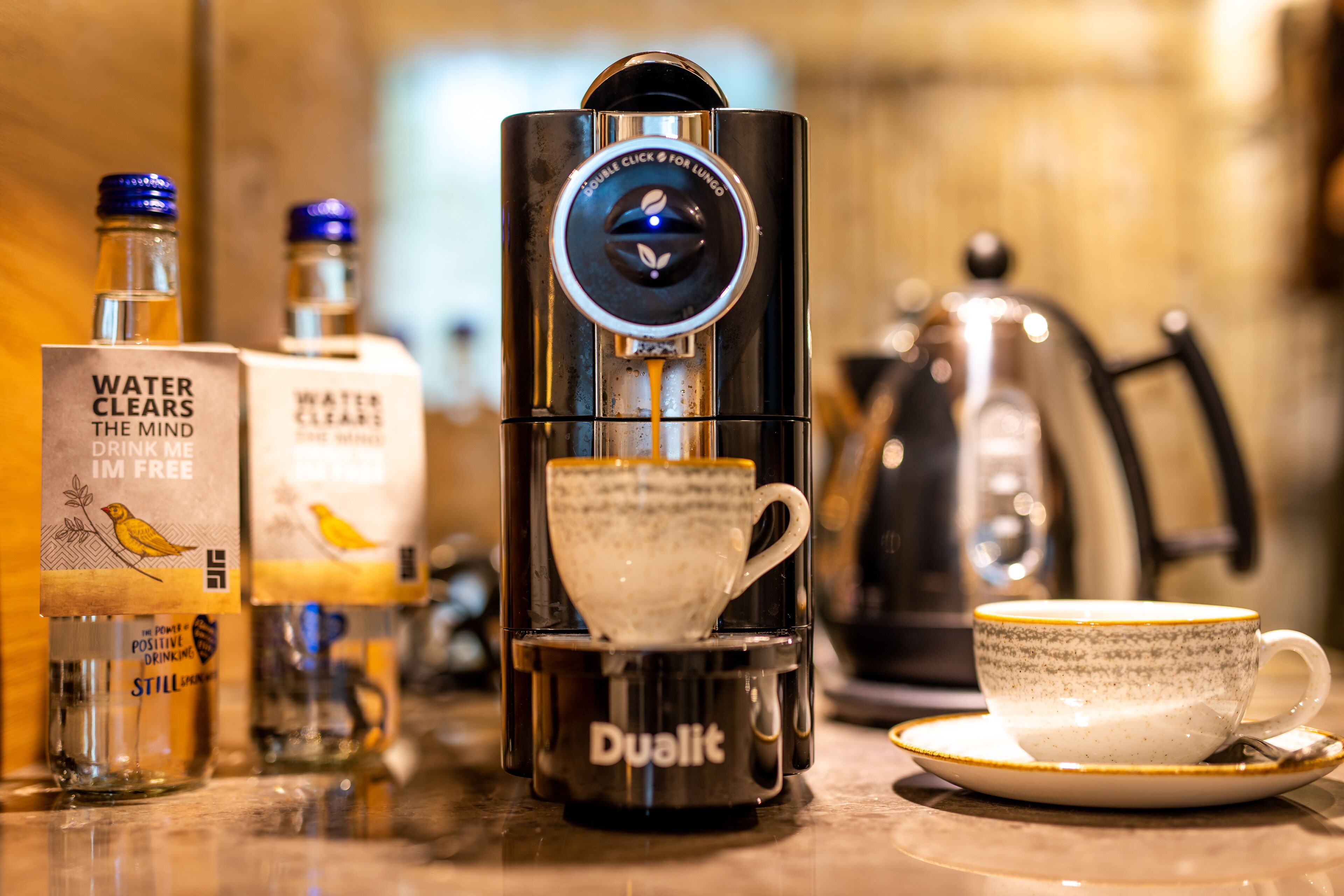 A coffee machine with a cup placed on its base, a coffee cup on a saucer, and two bottles of water on the counter.