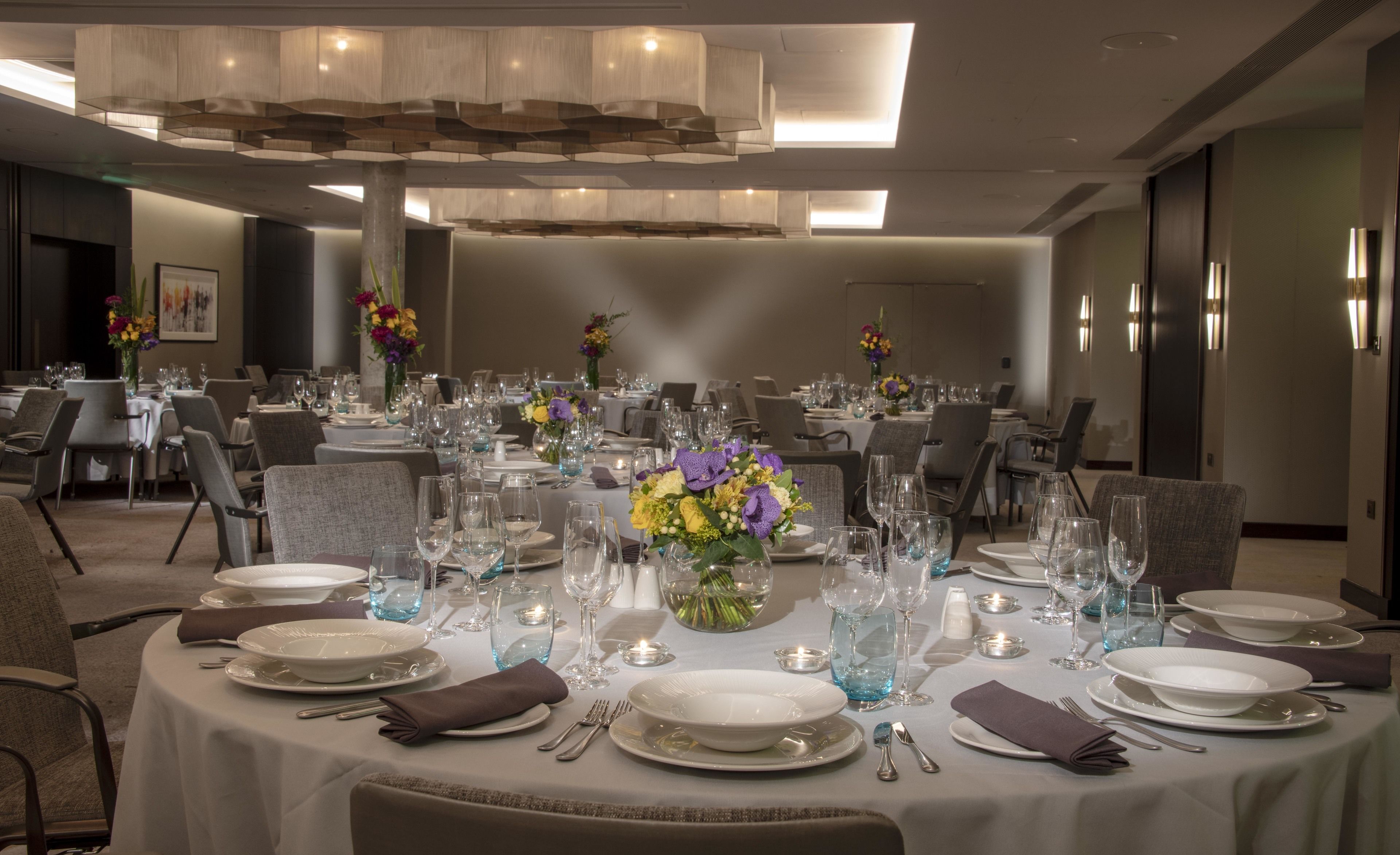 Une salle de banquet avec des tables rondes dressées pour le repas, agrémentées de nappes blanches, de verres bleus et de centres de table ornés de fleurs.