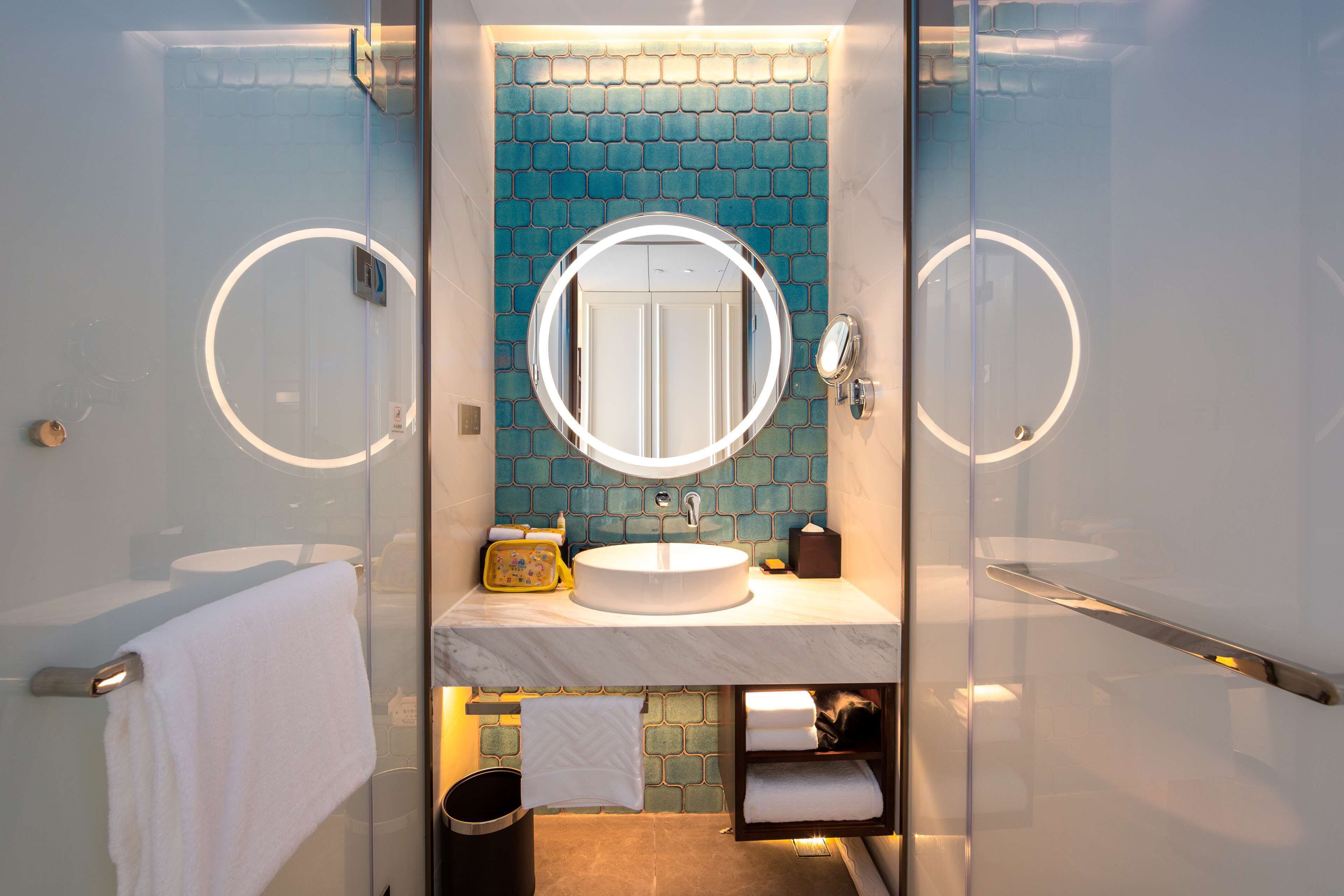 A modern bathroom with a glass-enclosed shower, a round mirror, a white sink, and a blue-tiled backsplash.