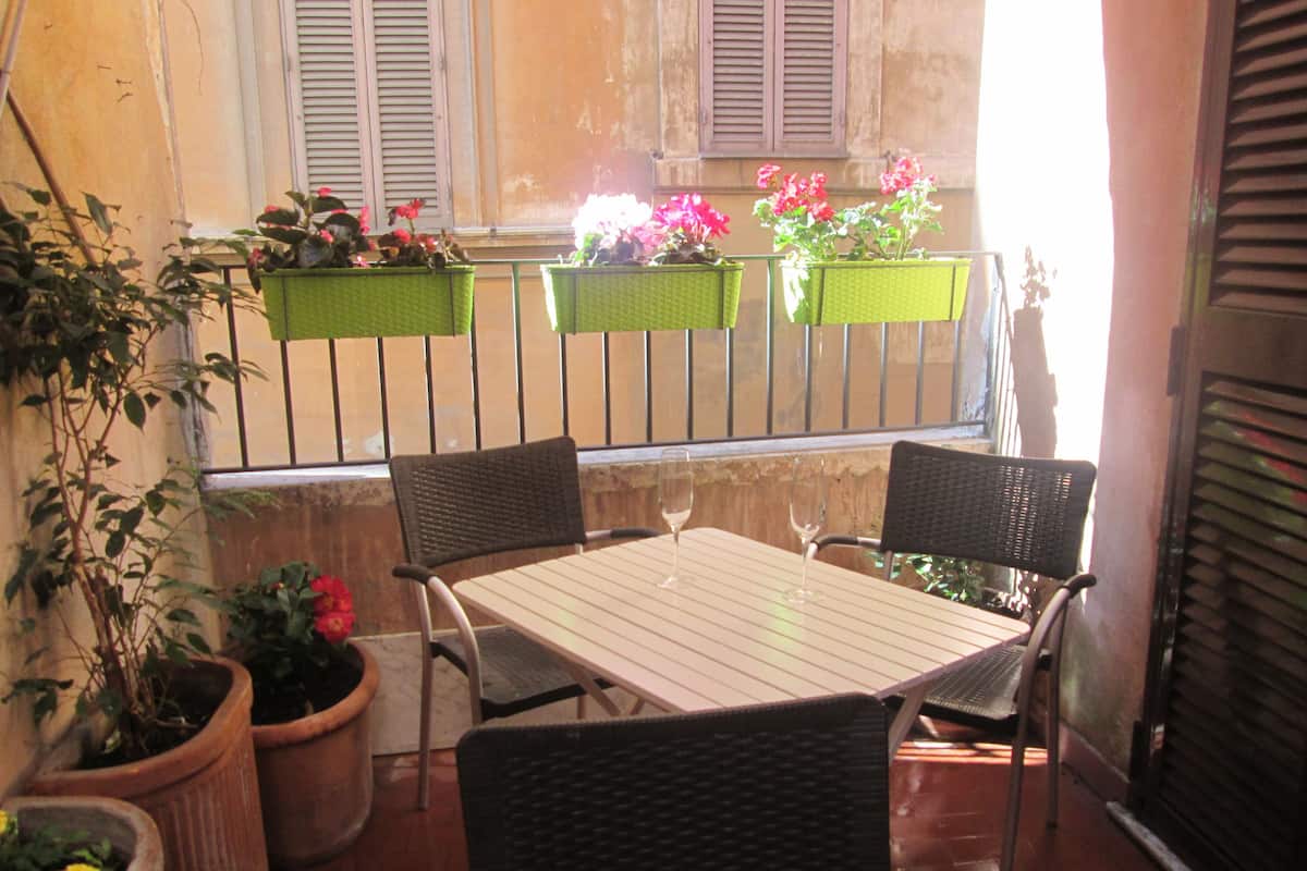 A balcony with a table and two chairs, potted plants, and flower boxes with pink flowers.