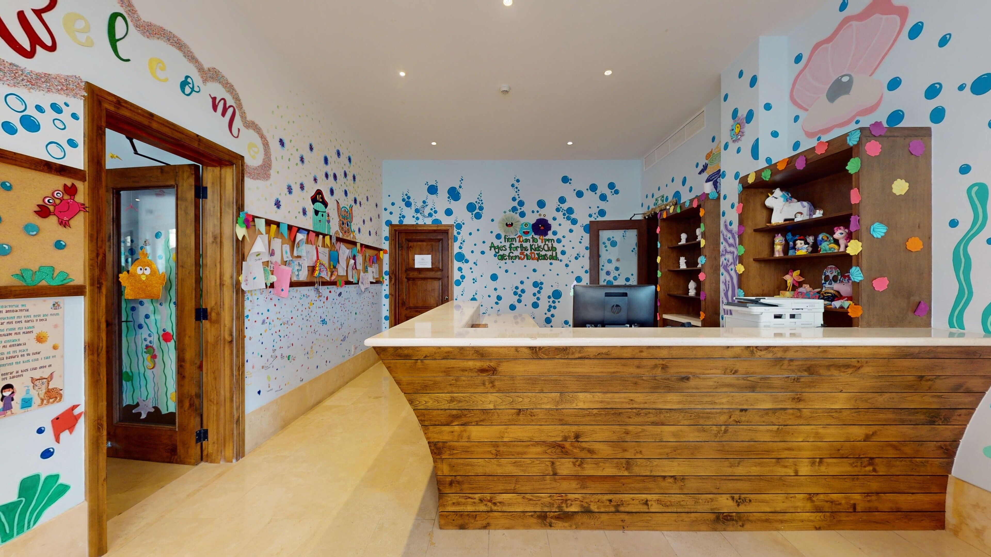 Een receptie met een houten balie, kleurrijke wanddecoraties en een bordje voor de kinderclub.