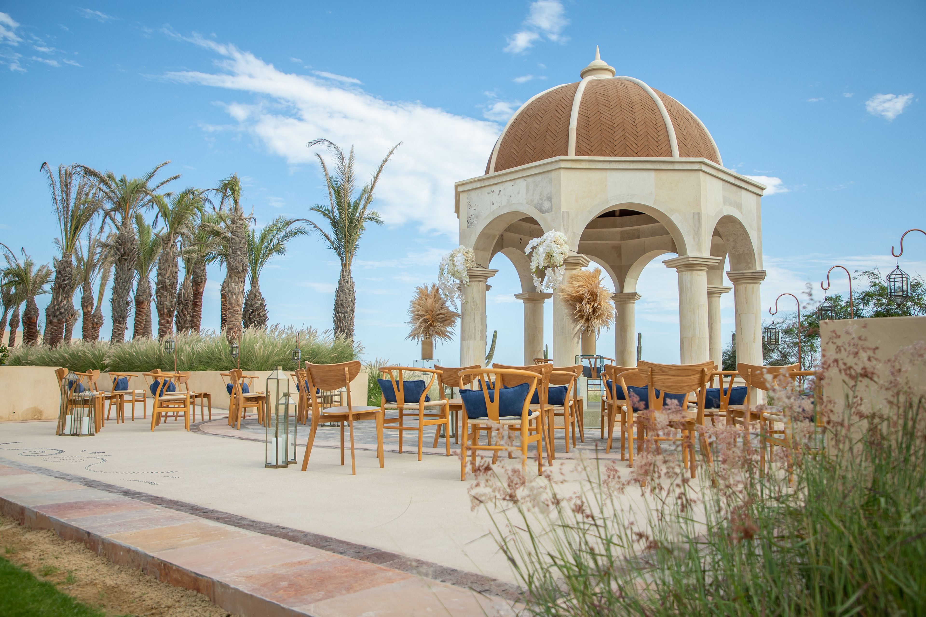 A gazebo with a dome roof and arches, surrounded by wooden chairs and tables, palm trees, and a clear sky.