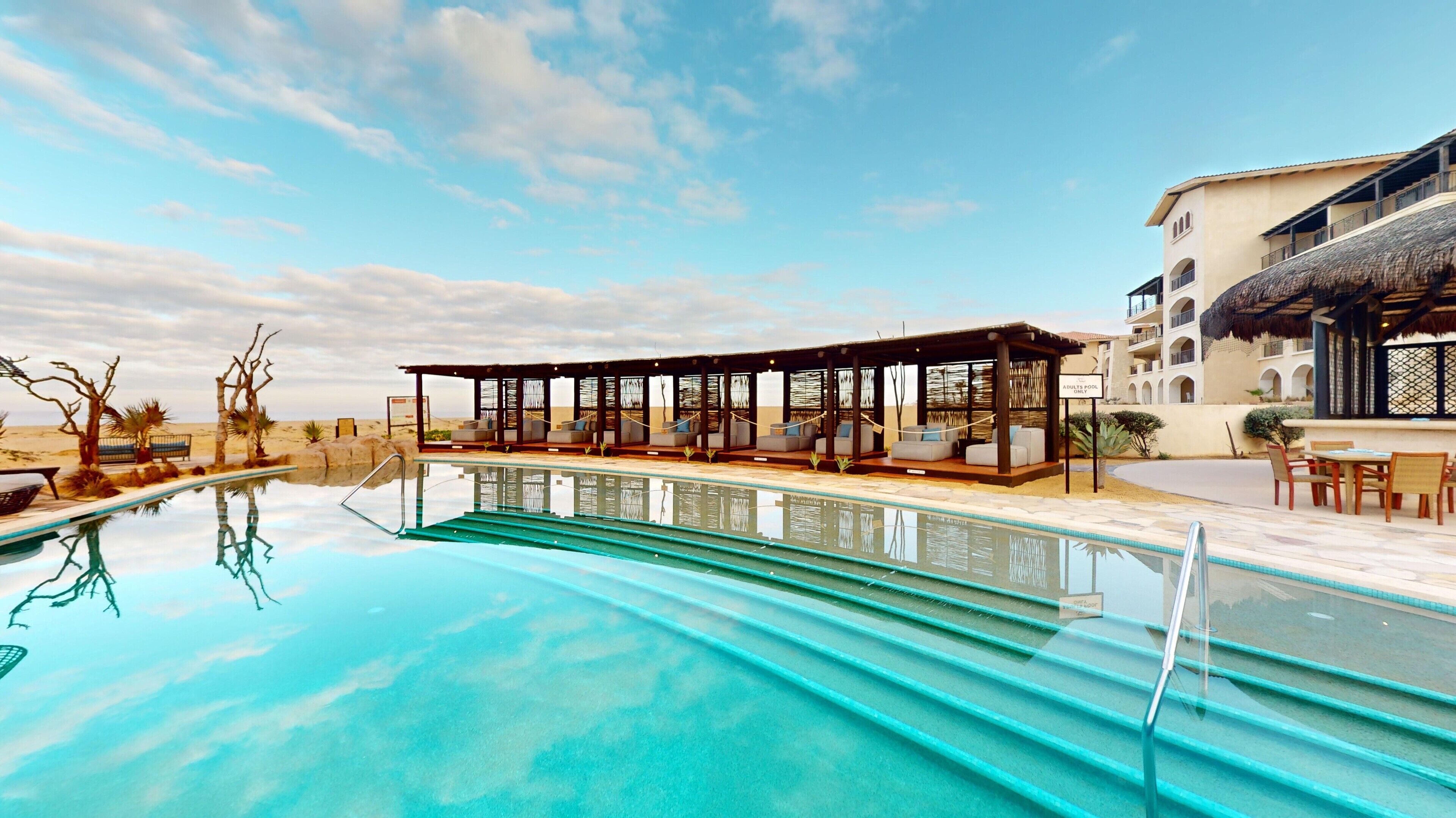 Un espace aménagé au bord de la piscine, comprenant un coin salon couvert, une piscine et un bâtiment doté d’un toit de chaume.