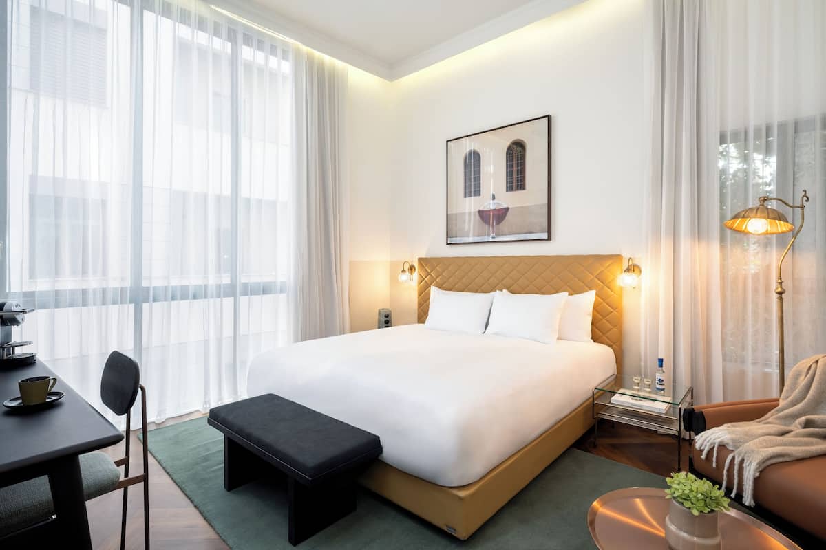 A hotel room with a large bed, a desk with a chair, a bench, a sofa, and a bedside table with a lamp.
