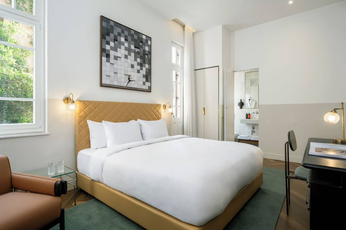 A modern hotel room with a large bed, a desk, a chair, a glass table, and a large window.