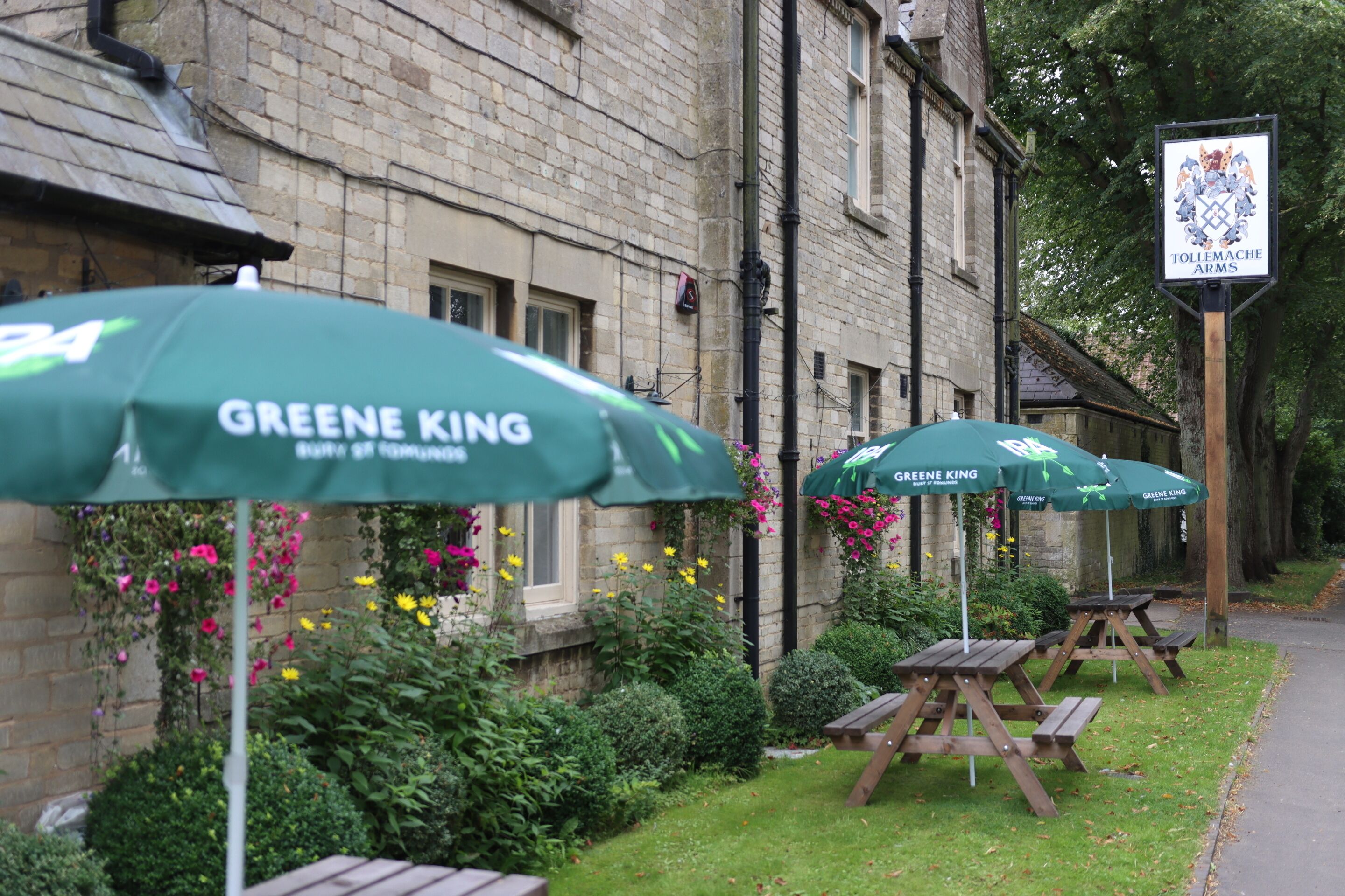 A building with a sign reading "TOLLEMACHE ARMS" and outdoor seating with "GREENE KING" umbrellas.