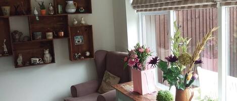 Una sala de estar acogedora con un sofá rosa, estanterías de madera con varios objetos decorativos y un ventanal con persiana a rayas.