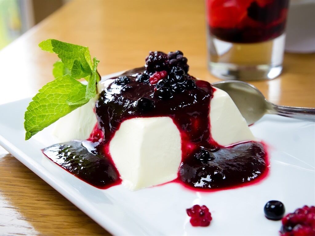Panna cotta servie avec une compote de fruits rouges et décorée de feuilles de menthe, le tout présenté sur une assiette blanche.