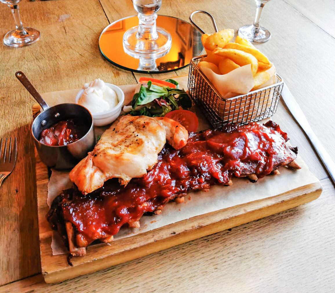 A wooden table with a plate of ribs, a basket of fries, a small bowl of sauce, and a glass of wine.