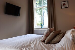 A neatly made bed with white linens and several pillows, a flat-screen TV mounted on the wall, a window with curtains, and a framed picture on the wall. - Tollemache Arms (Grantham)