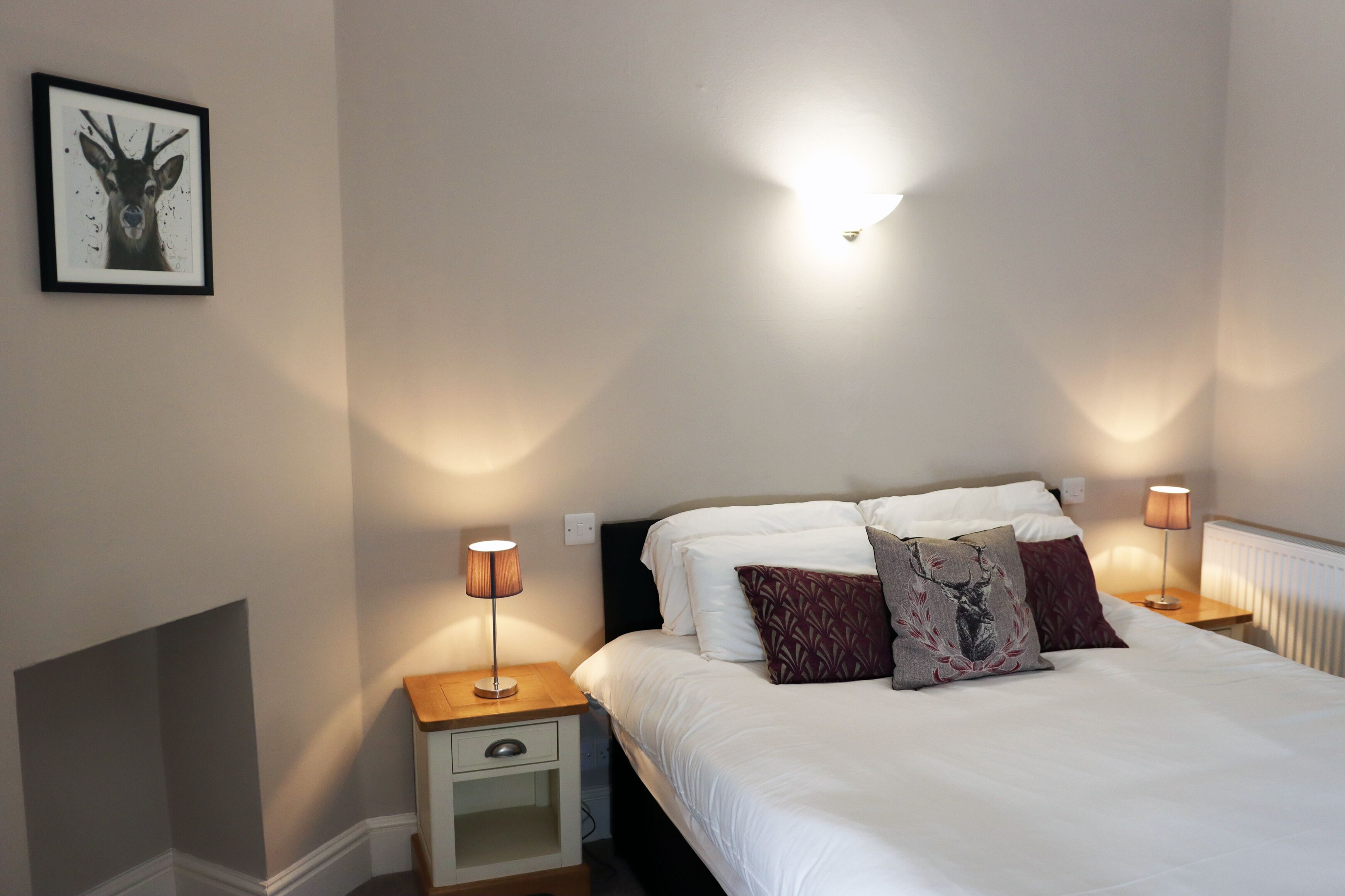 A bedroom with a bed, two bedside tables with lamps, a nightstand with a drawer, a framed picture on the wall, and a wall-mounted light fixture.