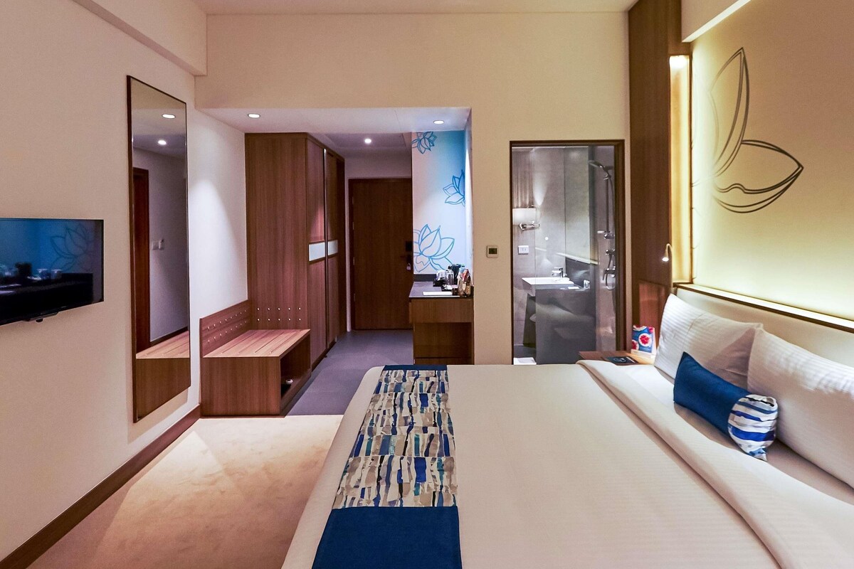 A modern hotel room with a large bed, a TV, a wardrobe, and a bathroom with a glass shower.