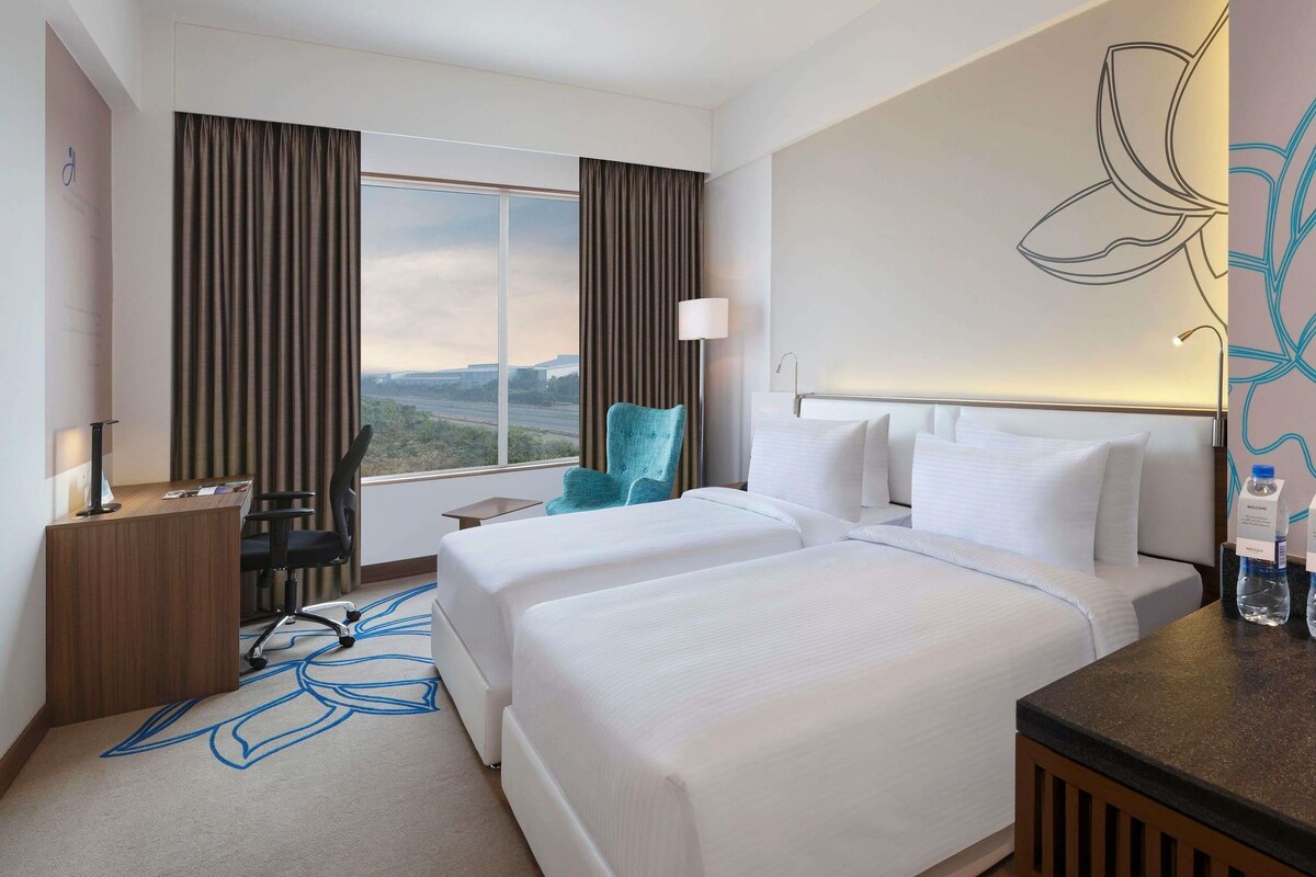 A hotel room with two beds, a desk, a chair, a lamp, and a view of the landscape.