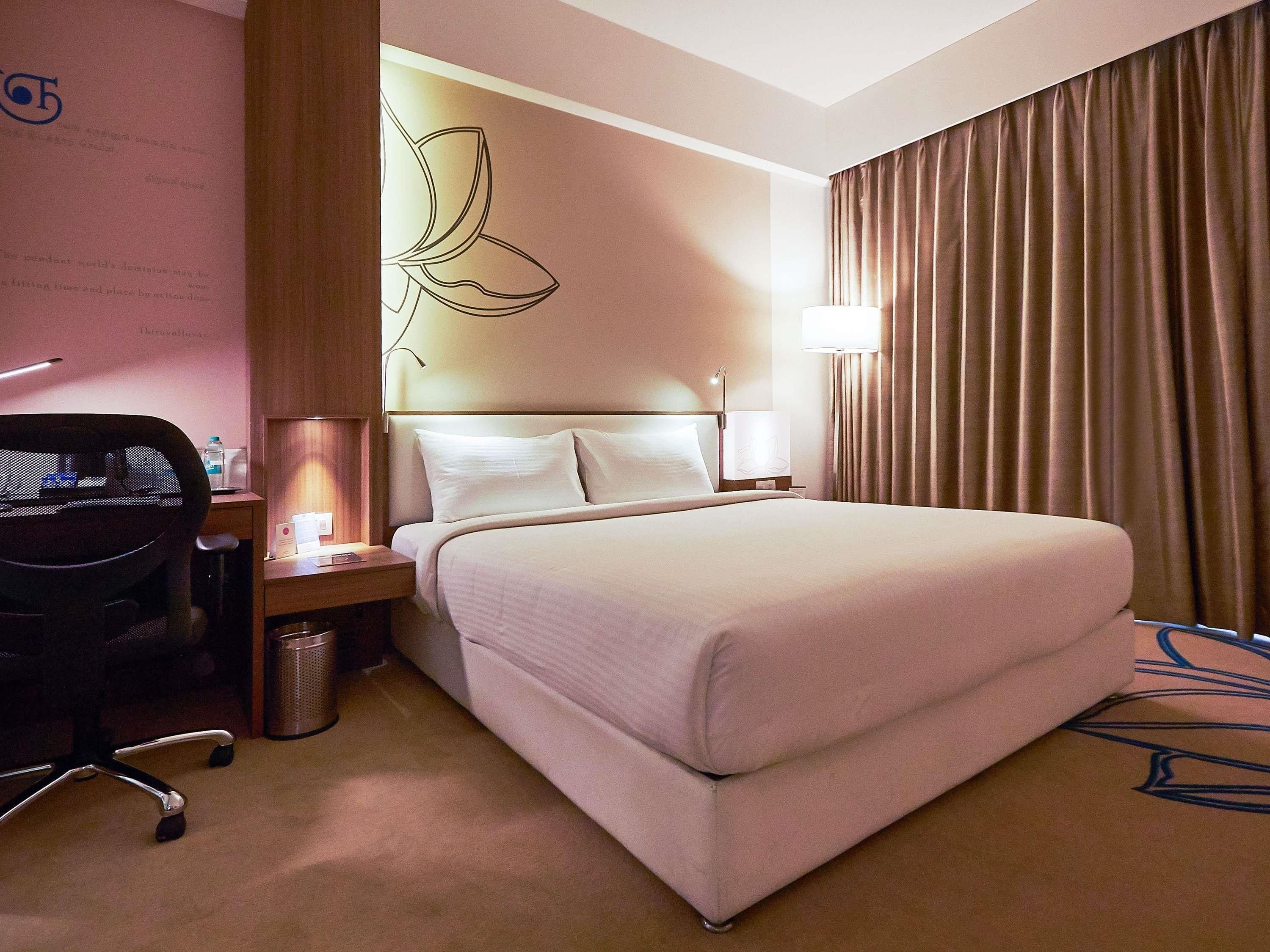 A hotel room with a bed, a desk with a chair, a lamp, a trash bin, and a wall with a logo.