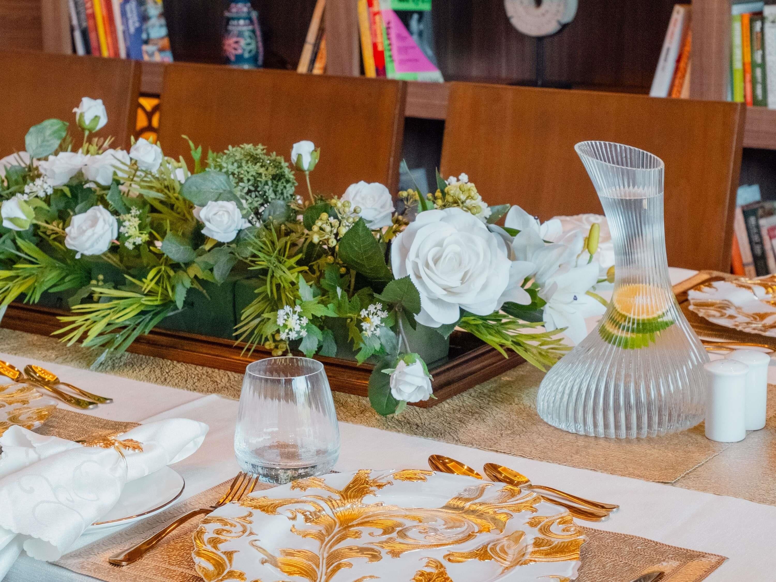 A table set with white tablecloths, gold leaf designs, and a centerpiece of white flowers.