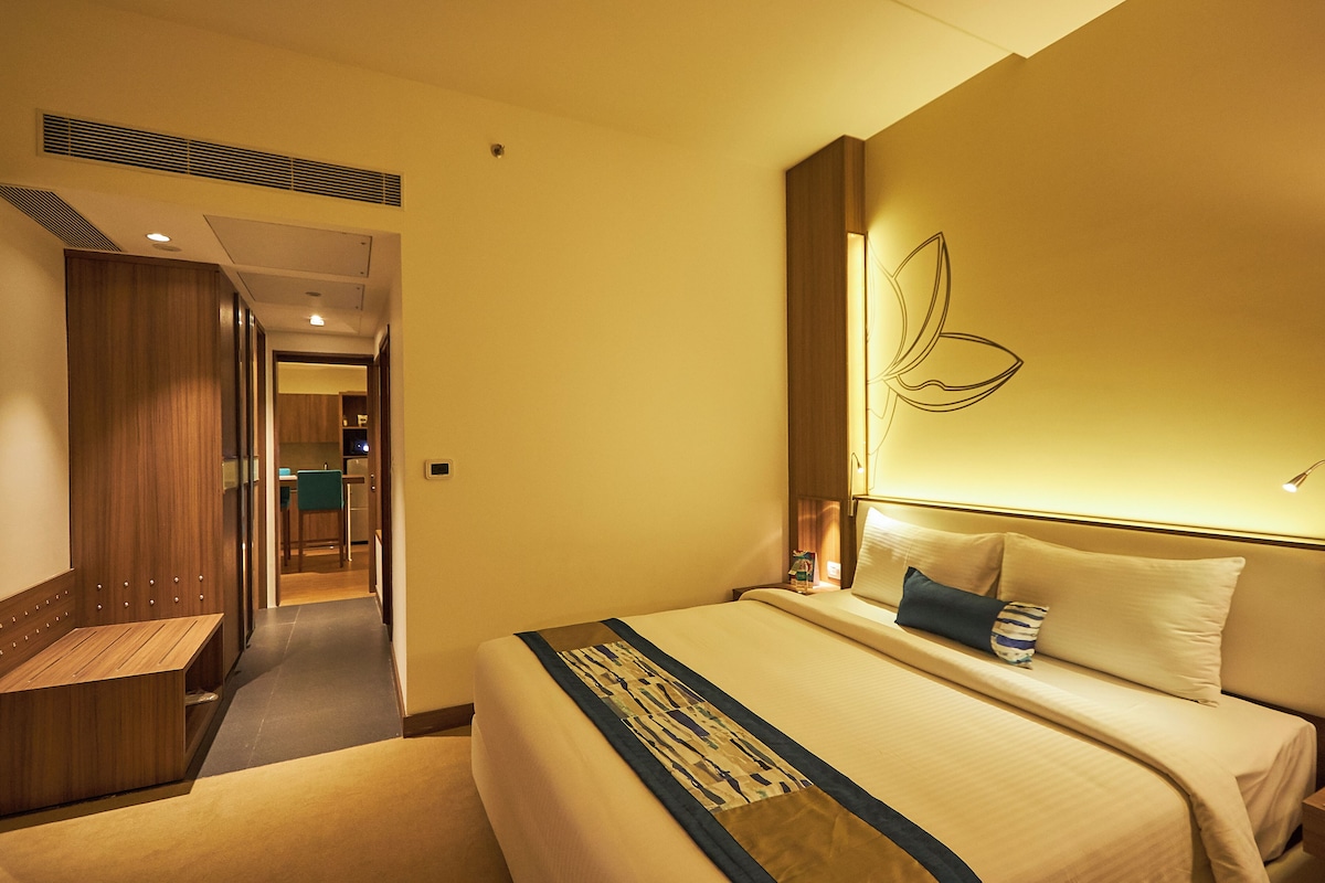 A hotel room with a bed, a wooden bench, a wall with a leaf design, and a hallway leading to a dining area.