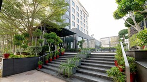 Un edificio moderno con una escalera que conduce a un acceso cubierto, rodeado de plantas en macetas y árboles.