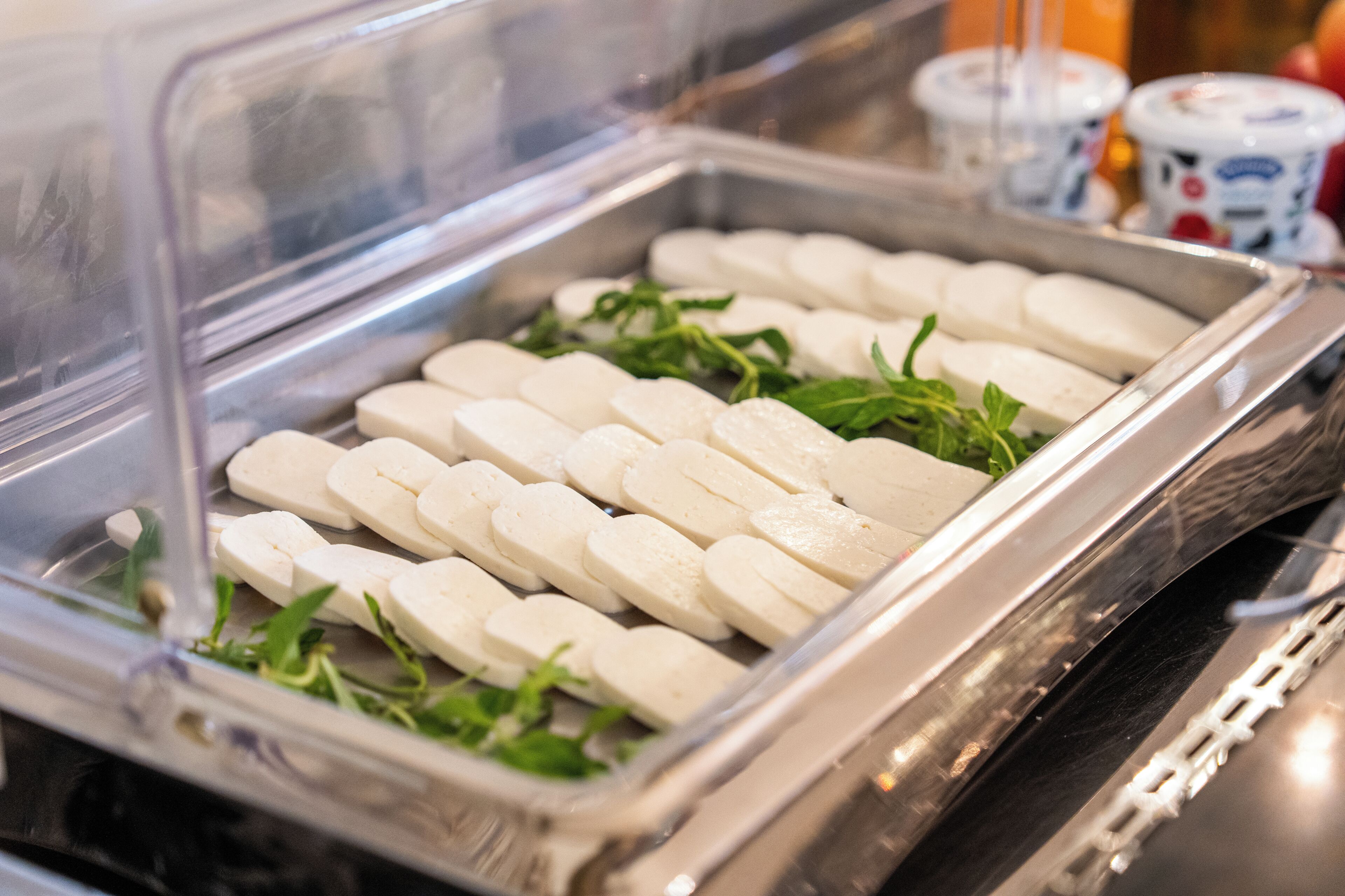 A refrigerated display case with sliced white cheese and green herbs.