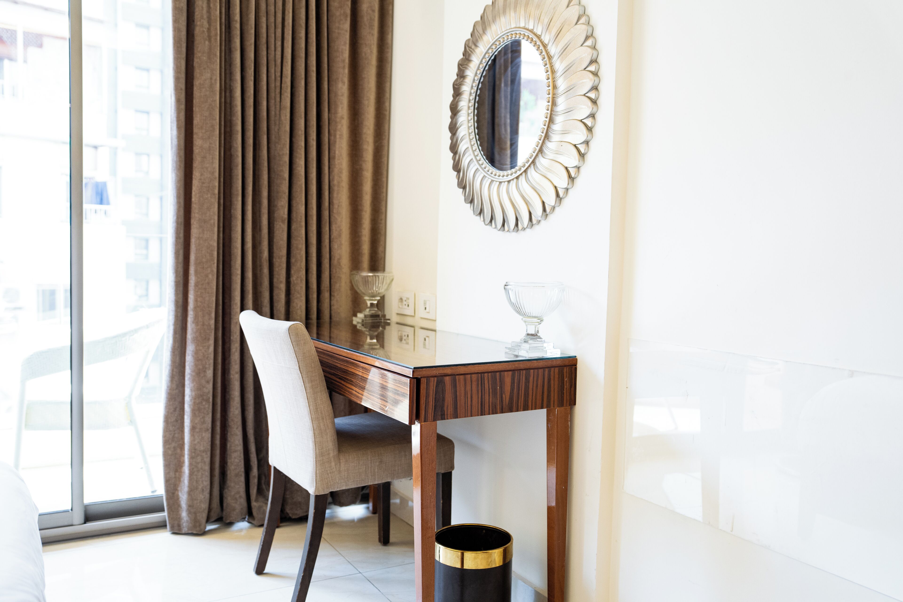 Une chambre d’hôtel avec un bureau en bois, une chaise, un vase en verre, un miroir décoratif et une fenêtre avec des rideaux.