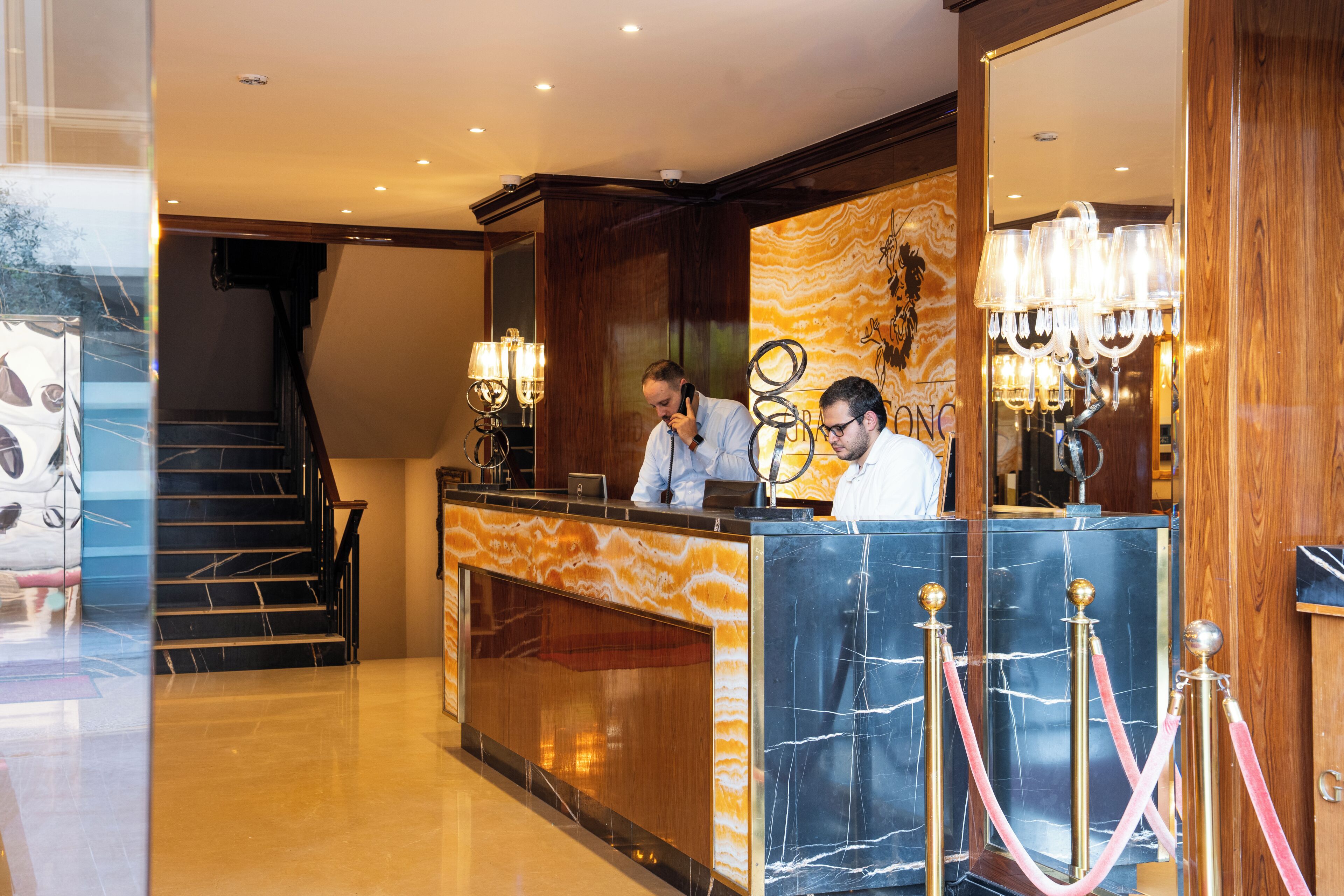 A hotel reception area with marble counters, a staircase, and a chandelier.