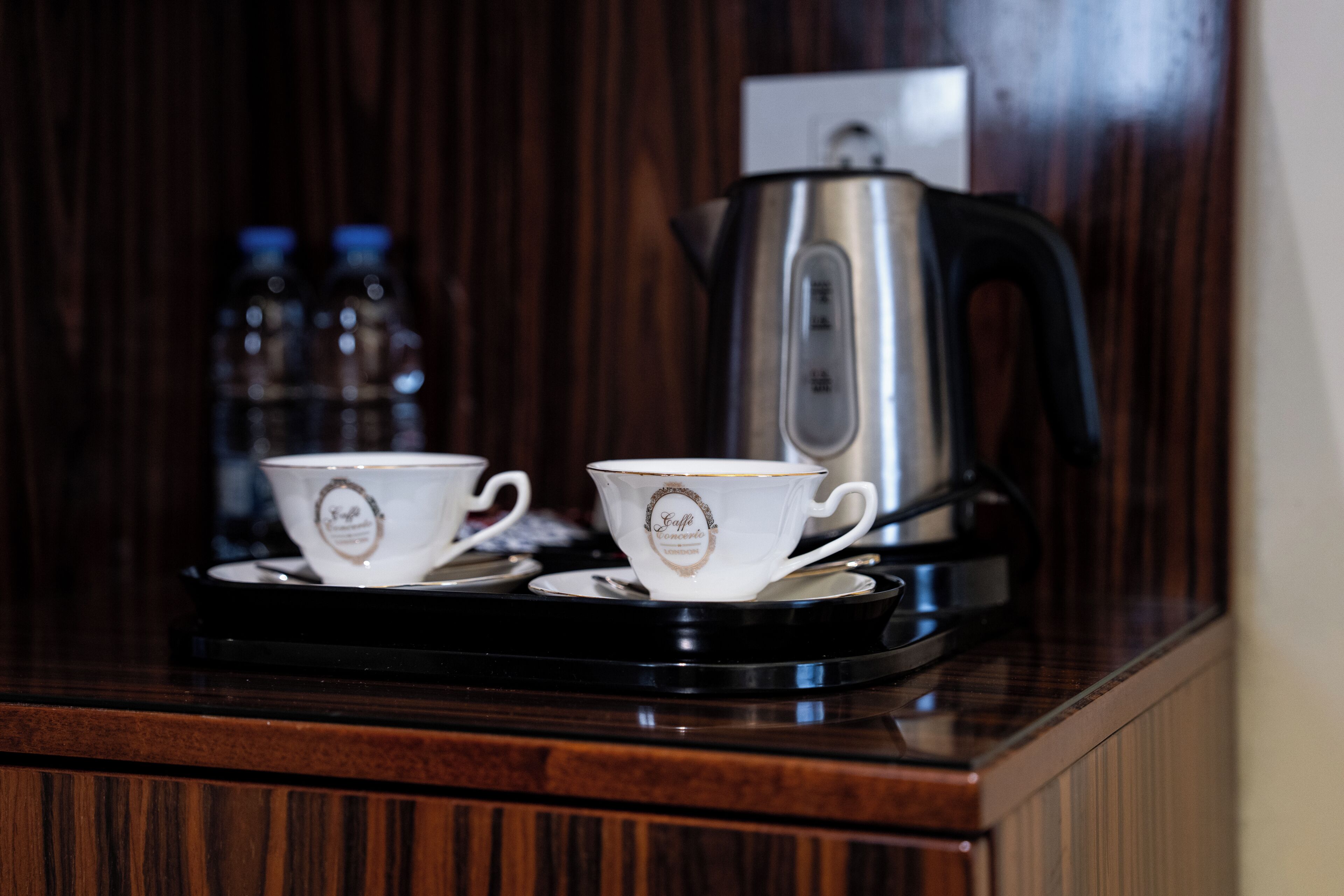 Two white cups with gold trim on a black tray, a silver coffee pot, and a wooden cabinet in the background.